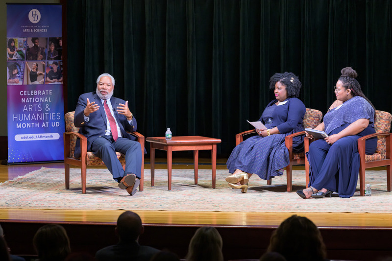 Three people on a stage inside a theater at the University of Delaware, seated in front of an audience, for a discussion about cultural heritage and preserving artifacts.